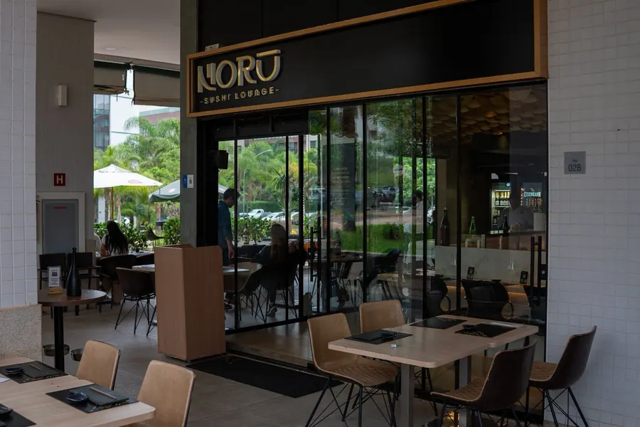A entrada do Norū de dia: o letreiro NORŪ SUSHI LOUNGE sobre as portas de vidro, mesas do lado de fora e palmeiras ao fundo.