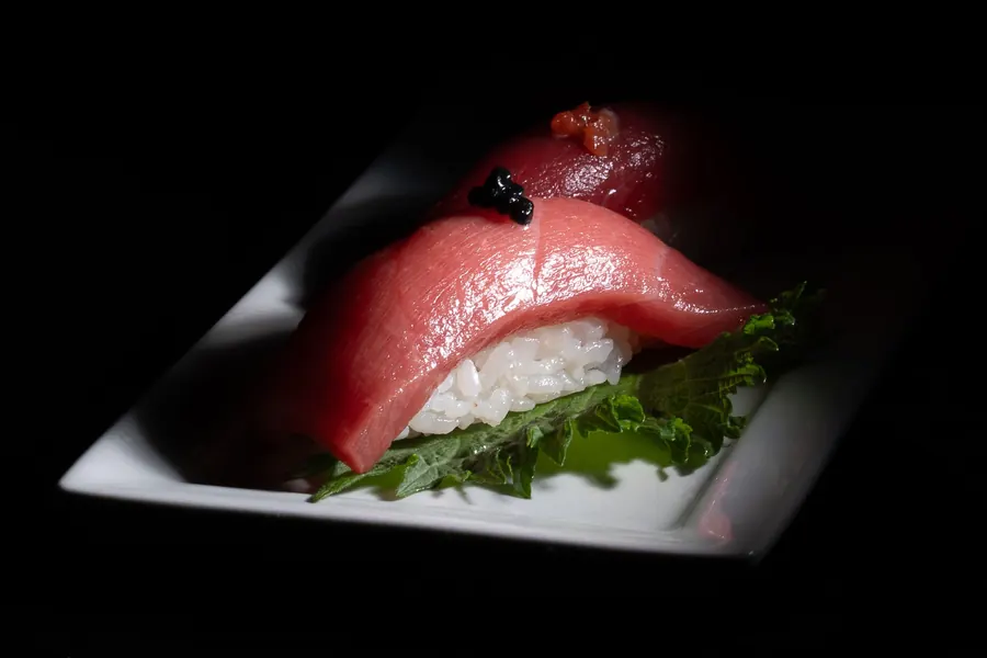 Dois niguiris de peixe vermelho sobre folha verde, um deles com ovas pretas por cima, em prato branco sobre fundo escuro.